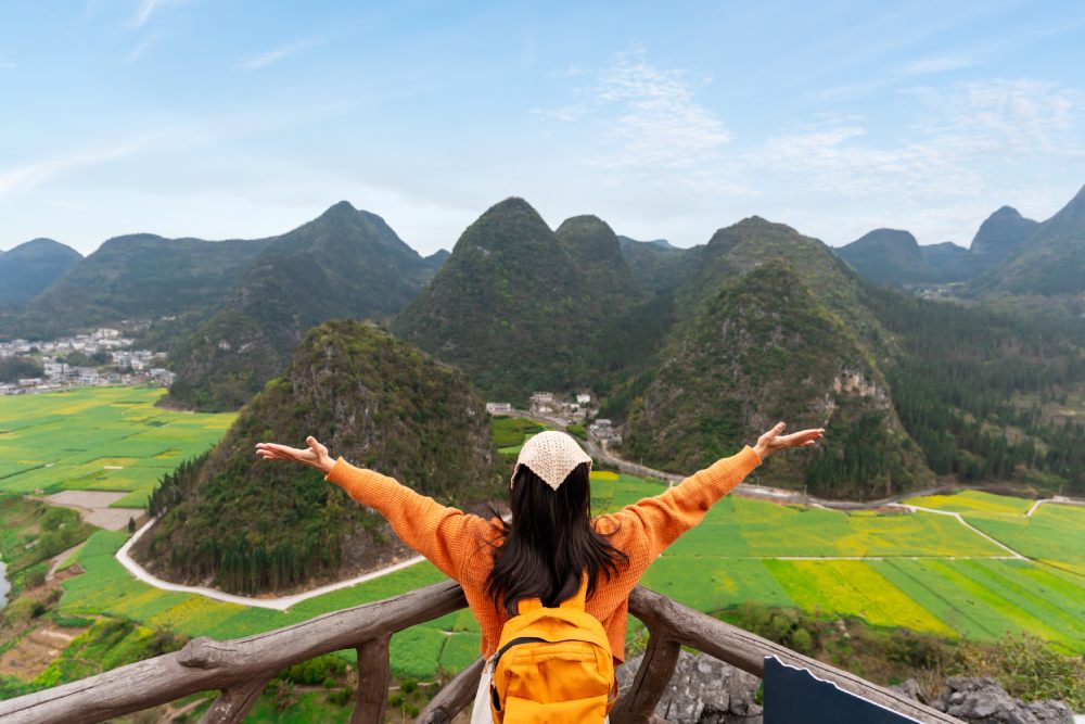 Foto de mujer viendo el panorama de las montañas Kársticas de China desde una terraza con acabado rustico. Esta con las manos abiertas y viendo el panorama natural.